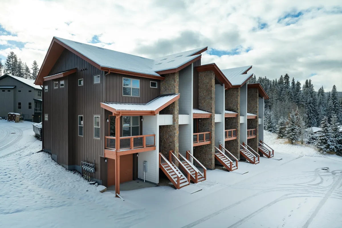 A modern architectural chalet with stone accents situated in a cozy, snow-covered landscape. The scene hints at après-ski relaxation, indoor warmth, and outdoor adventure, with the inviting glow of a warm interior contrasting with the winter surroundings.