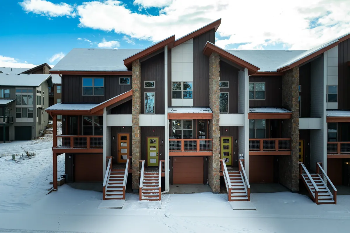 A beautiful chalet exterior featuring large windows, wooden stairs, stone accents, and a snow-covered landscape. The home appears inviting and private, nestled serenely in a wintry natural setting, hinting at nearby skiing and winter activities.
