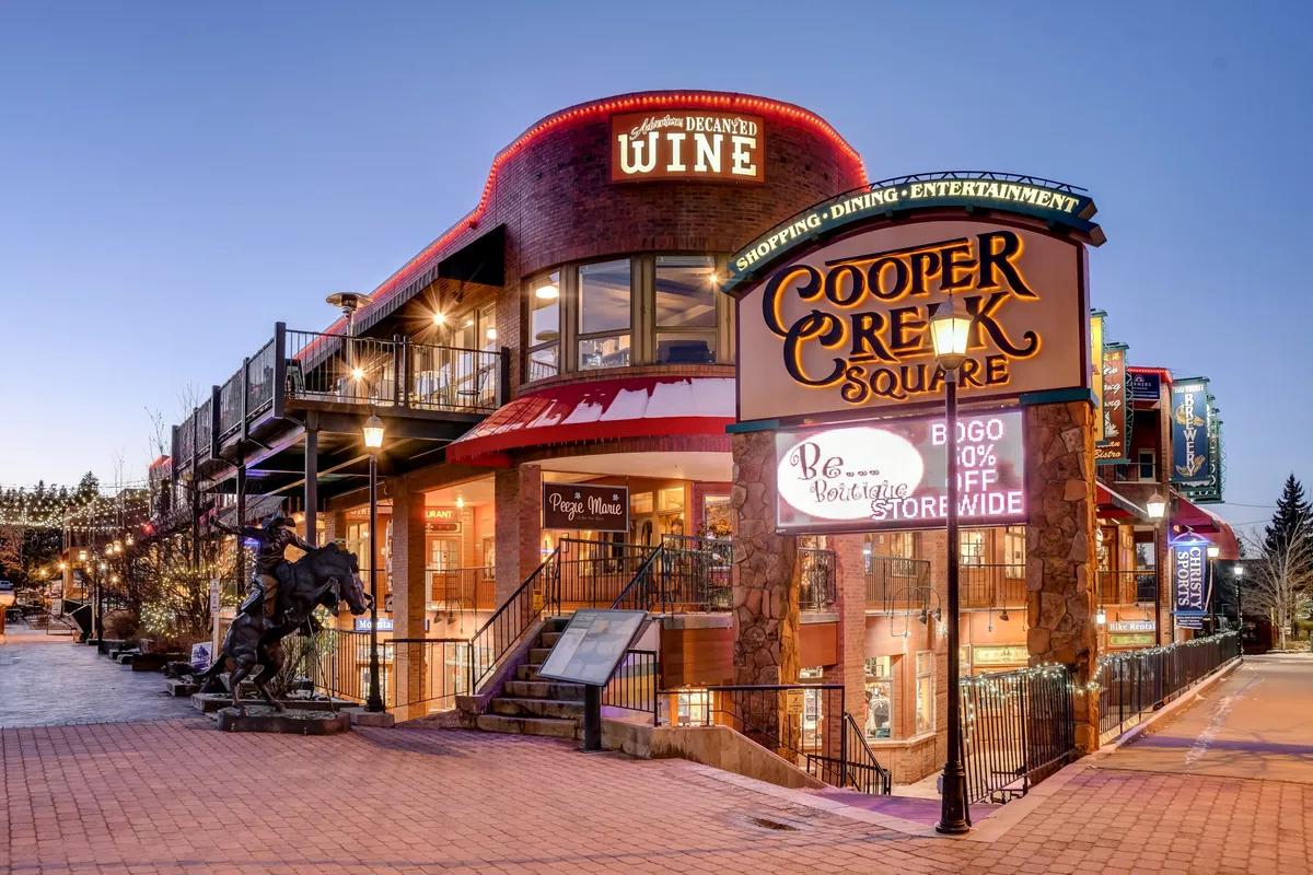 The vibrant Cooper Creek Square, featuring its iconic sign, festive decorative lighting, inviting outdoor seating, and a horse sculpture. The scene hints at a bustling community hub ideal for shopping, dining, and entertainment within walking distance of the property.