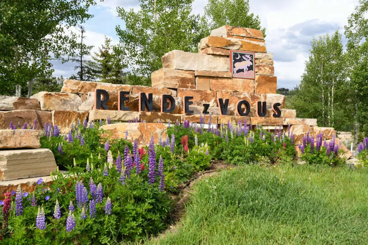 A beautifully landscaped entrance with a stone sign reading 'RENDEZ-VOUS,' surrounded by colorful flowers, green grass, and mature trees. The scene conveys the inviting atmosphere of the Rendezvous neighborhood and highlights the area's natural beauty and community feel.