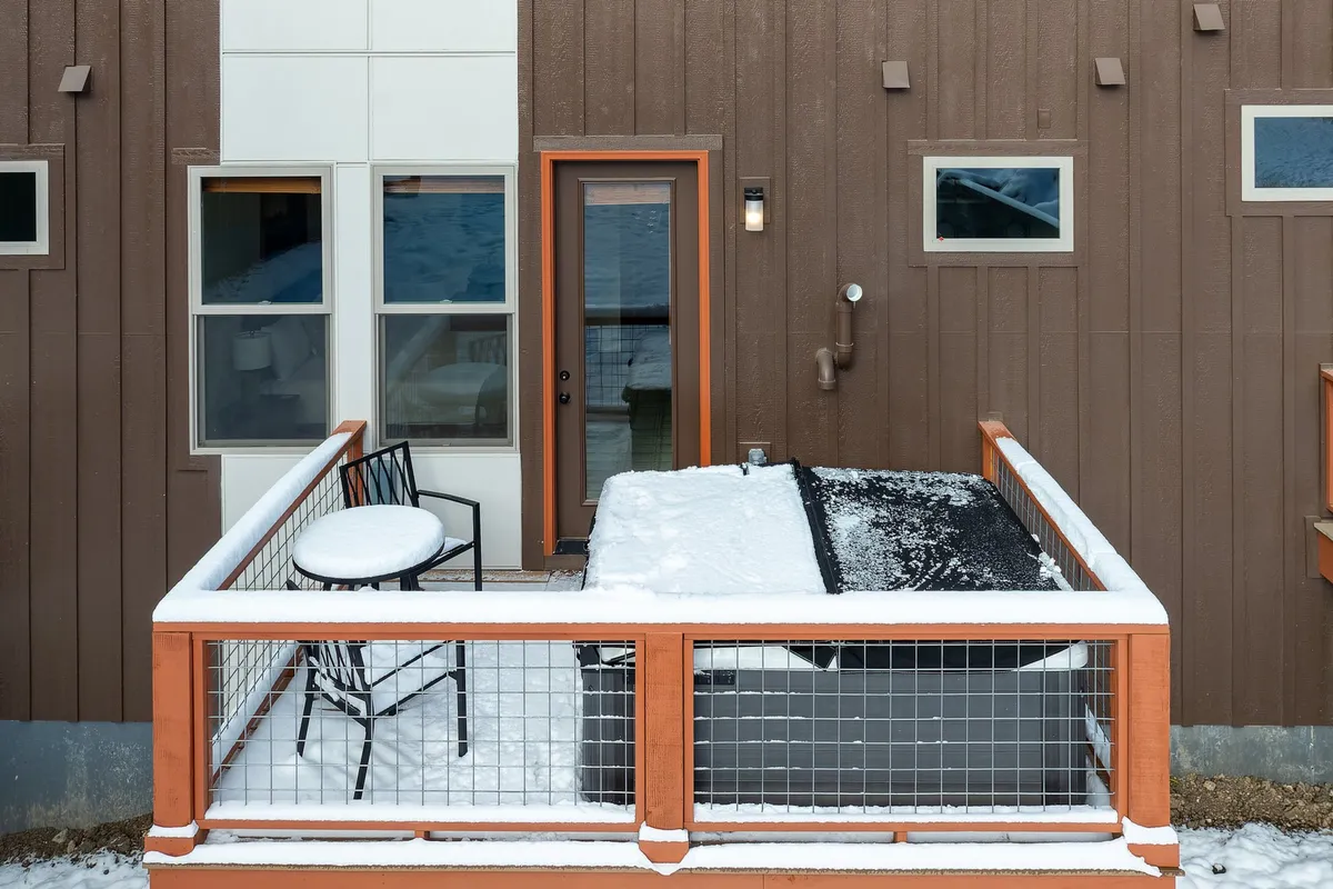 A cozy outdoor hot tub and seating area set against a snowy backdrop, with inviting steam rising and comfortable furnishings suggesting post-adventure relaxation. The entrance door and surrounding snowy landscape make clear the seamless transition from outdoor excitement to private comfort.