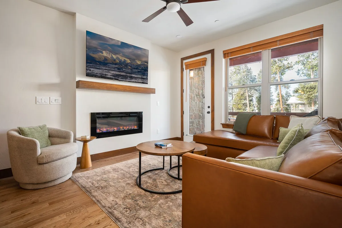 A sunlit living room anchored by a modern electric fireplace, with a leather sofa, wooden coffee table, and tasteful artwork on the wall. The space is bright and open, designed for relaxation and socializing, and it exudes both warmth and contemporary mountain charm.